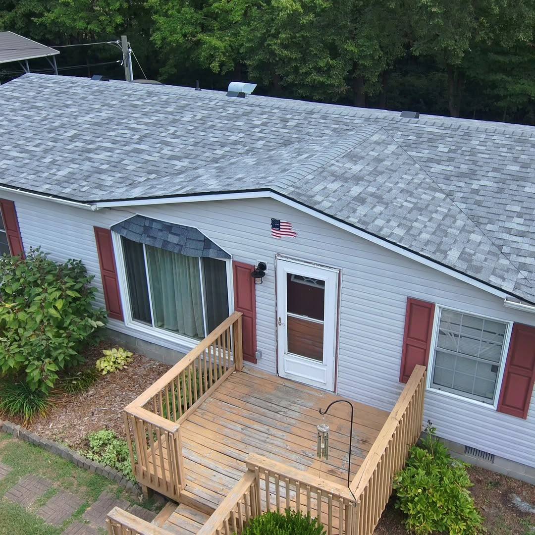 A ranch house photographed from above, its new grey architectural shingle roof running the width of the frame over white lap siding, burgundy shutters and a wooden deck at the front door.