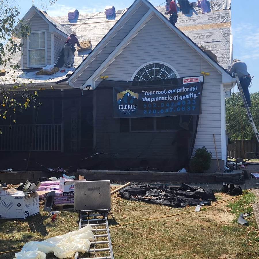 A white house mid-roof-replacement with the old shingles stripped away, protective sheeting draped down the wall and a company banner hung across it, tools and debris on the lawn below.