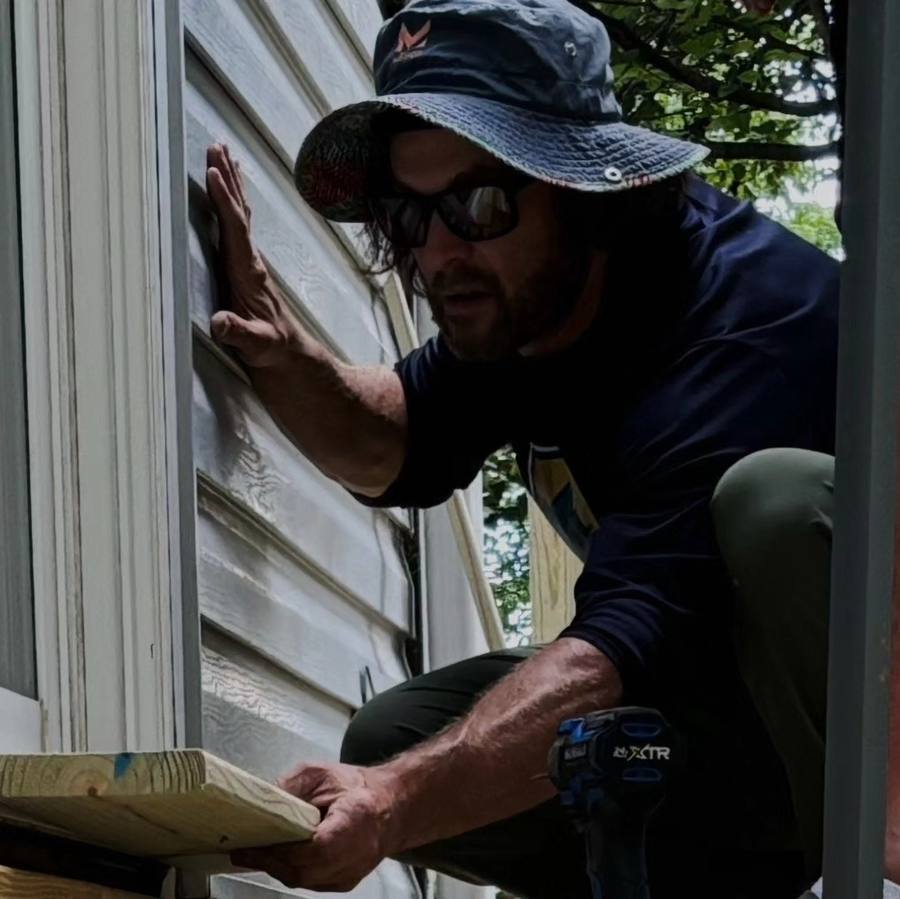 A worker in a wide-brimmed hat and sunglasses holding a length of vinyl siding flat against a wall as he sets it in place.