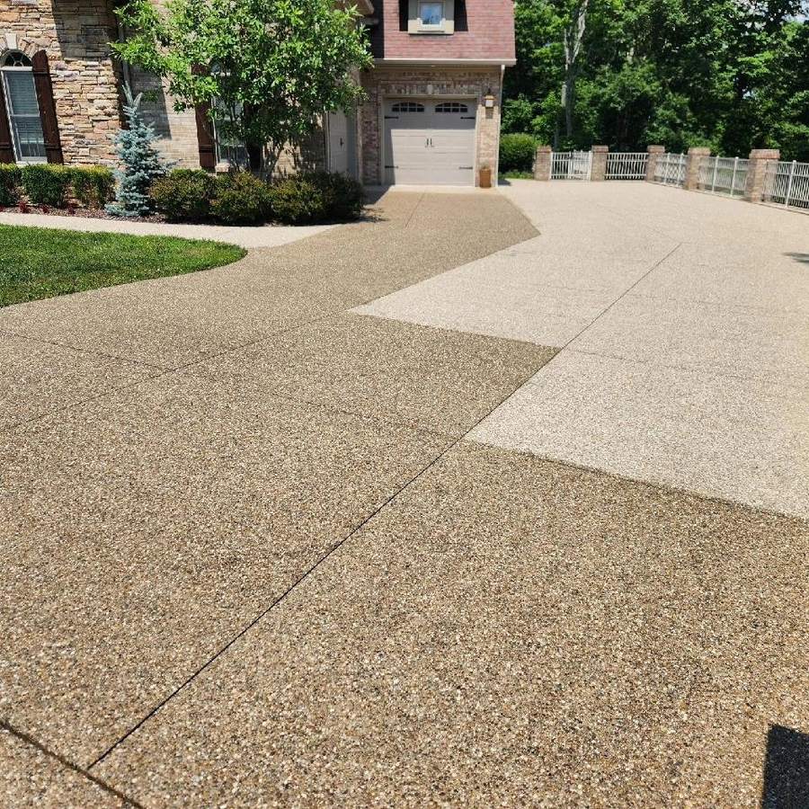 A wide exposed-aggregate concrete driveway with a smooth banded inlay running across it, leading up to a brick house with a garage.